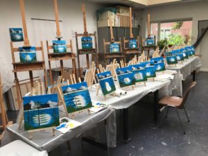 Een rij ezels en een rij tafels in het schilders atelier met Bob Ross schilderijen in wording